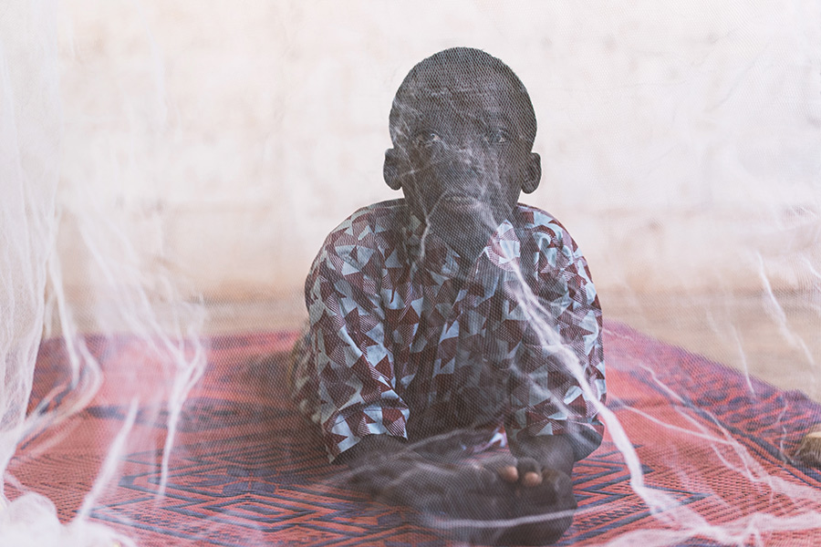 A young black boy wearing a colourful shirt is lying on a bed under a bed net. These are used to avoid getting bitten by mosquitoes, and can help reduce transmission of malaria. 